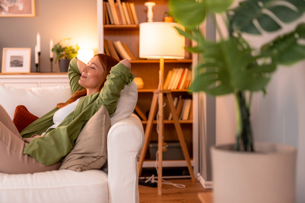 Person entspannt auf einer Couch in einem Wohnzimmer mit Bücherregal und Lampe im Hintergrund