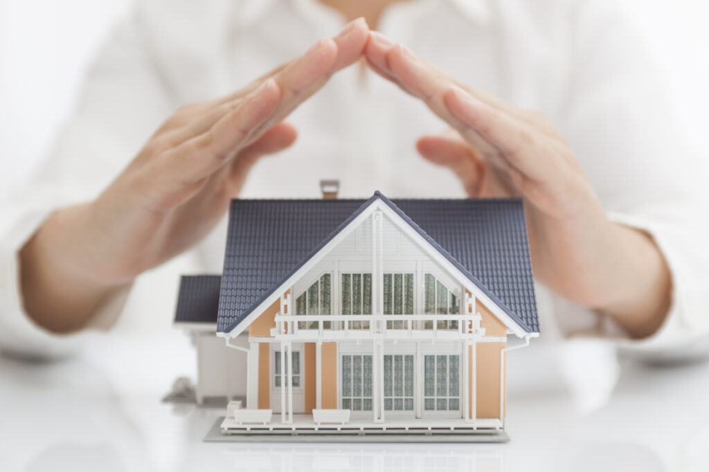 Modellhaus wird von schützenden Händen überdacht und symbolisiert Sicherheit für Wohneigentum