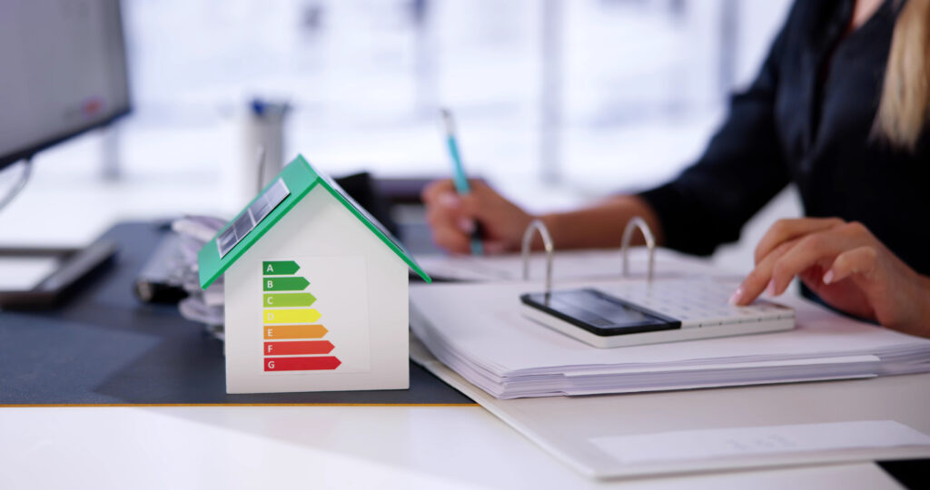 Modellhaus mit Energieeffizienzklassen A bis G auf dem Dach neben Unterlagen und Tablet auf einem Schreibtisch