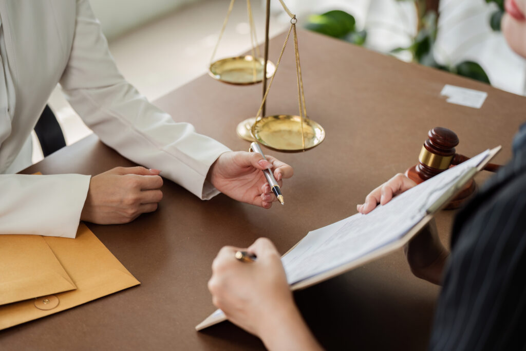 Eine Notarin im weißen Blazer zeigt einer weiteren Person am Tisch etwas auf einem Klemmbrett.