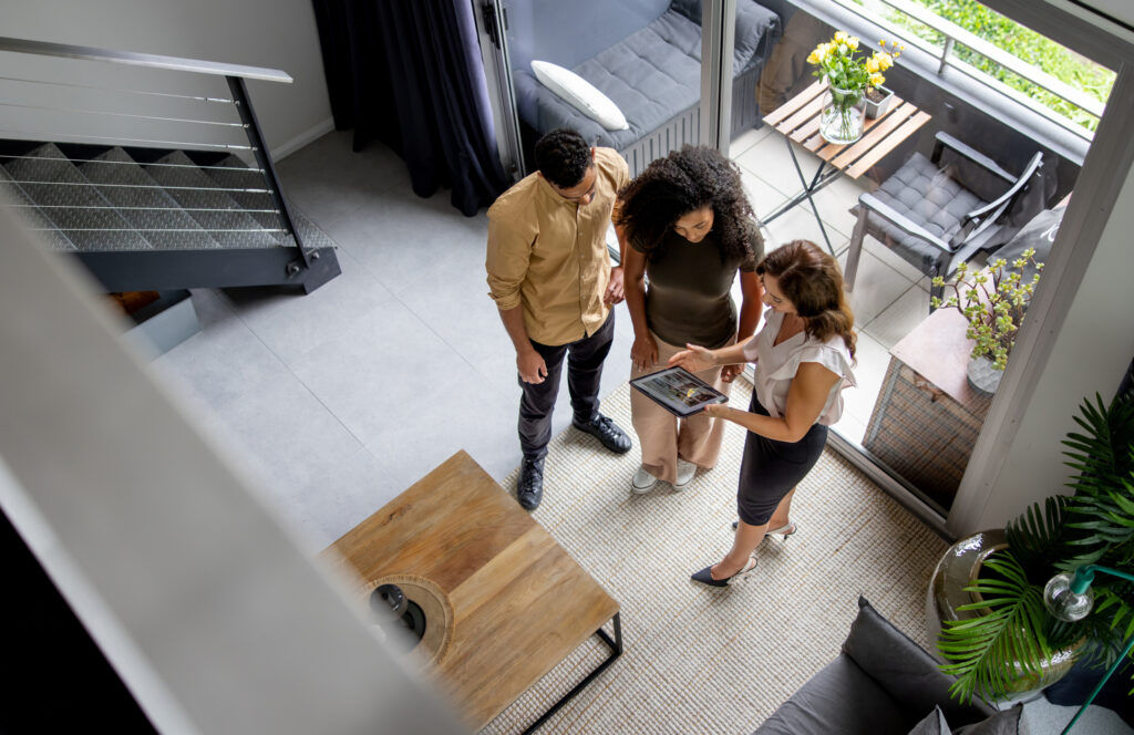 Immobilienmaklerin zeigt jungem Paar Grundriss auf Tablet.