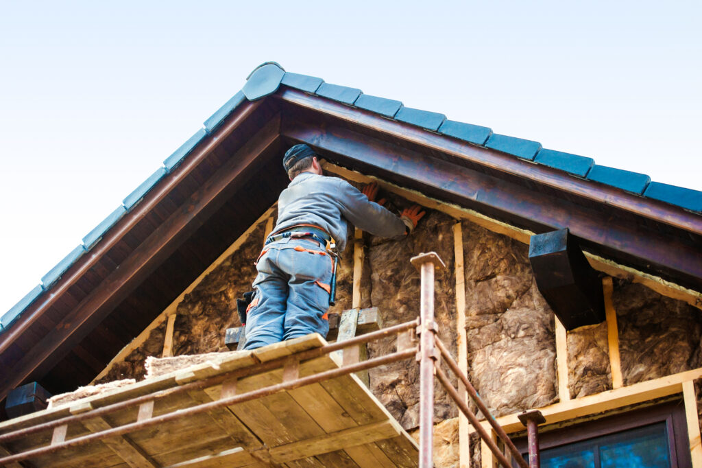 Arbeiter bringt Wärmedämmung an Hausfassade an, sichtbar sind Holzlatten und Dämmstoffe im Dachbereich